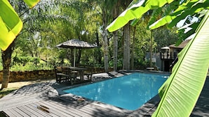 A swimming pool surrounded by a wooden deck, with a table and chairs under a shaded area, and lush greenery in the background. - BON Hotel Rustenburg (Rustenburg)