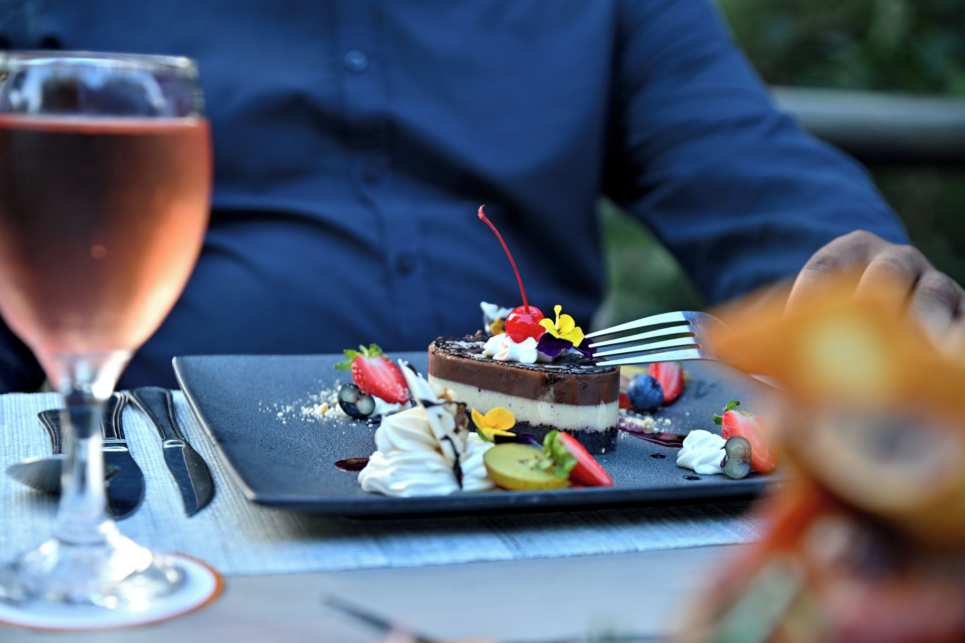 Un plato de postre con diversas frutas y una cereza encima, acompañado de un vaso de bebida rosa.