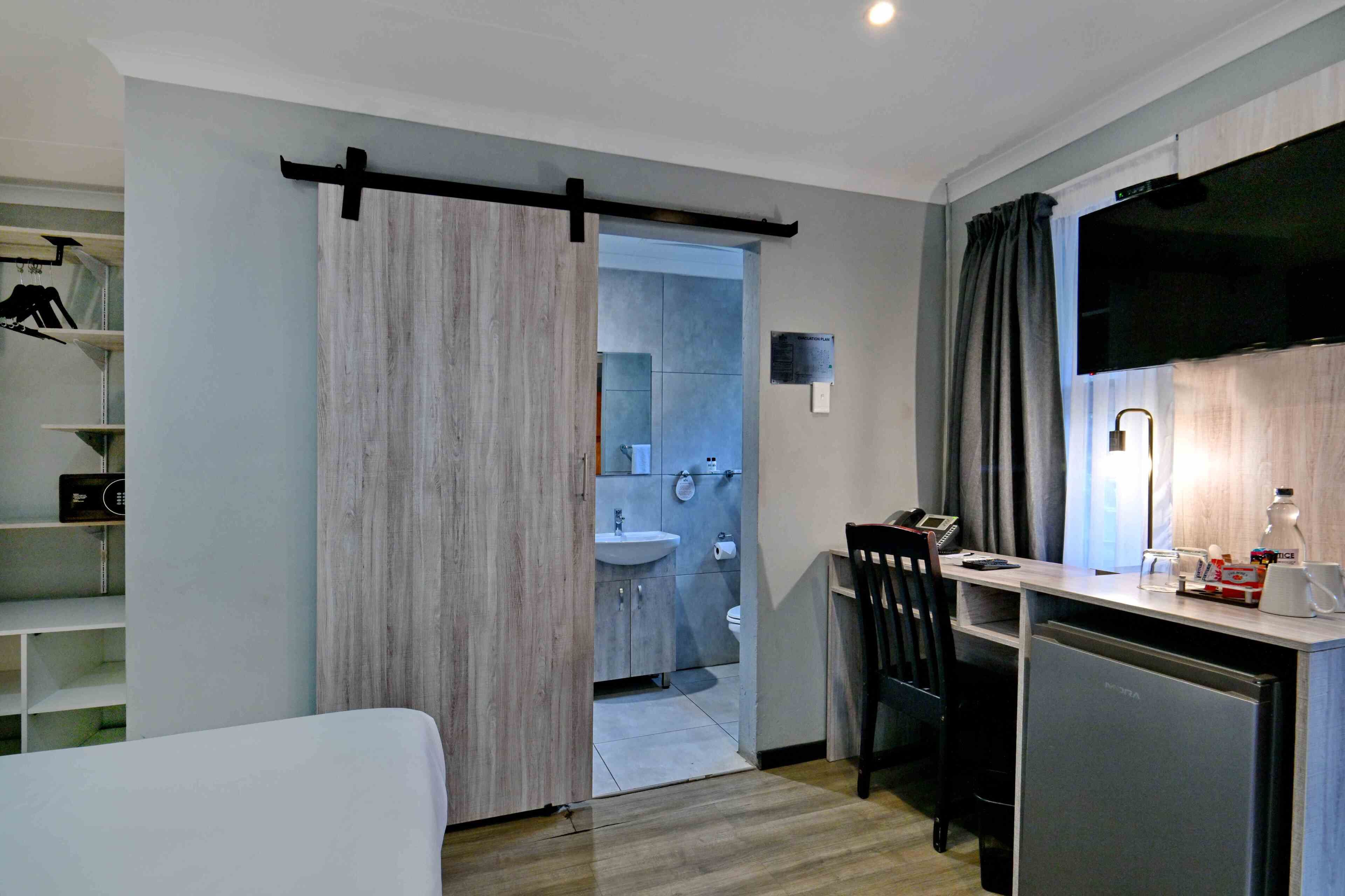 Een kleine hotelkamer met een kitchenette, een schuifdeur naar de badkamer, een bureau met stoel en een aan de muur bevestigde televisie.