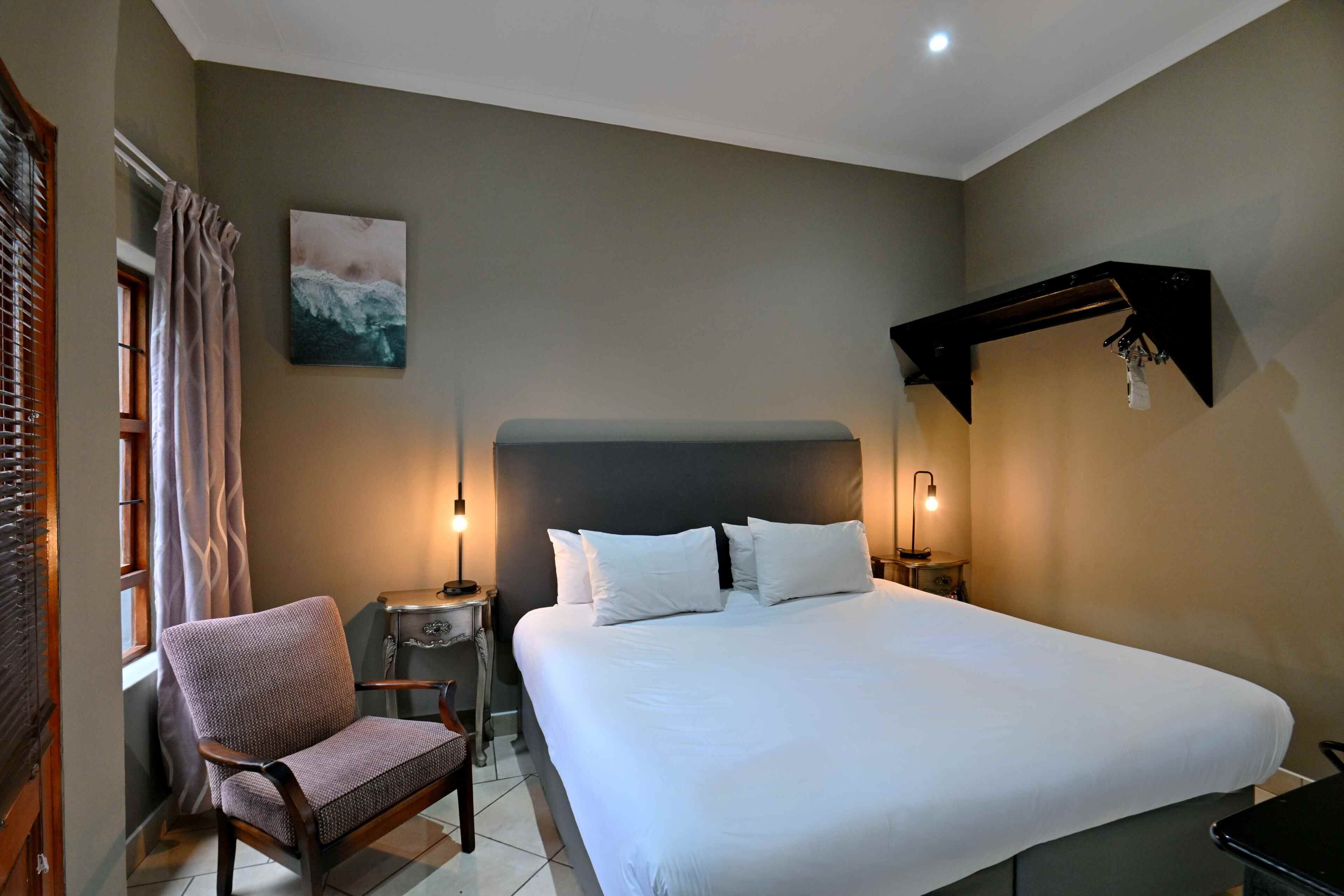 Une chambre à coucher avec un grand lit, une chaise, une table de chevet, une étagère fixée au mur et une fenêtre avec des rideaux.