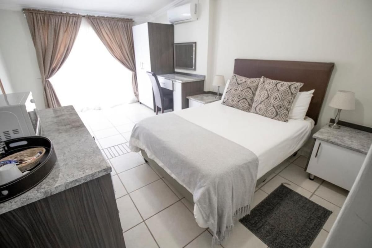 Quarto de hotel com cama, escrivaninha, cadeira, televisão e pequena cozinha.