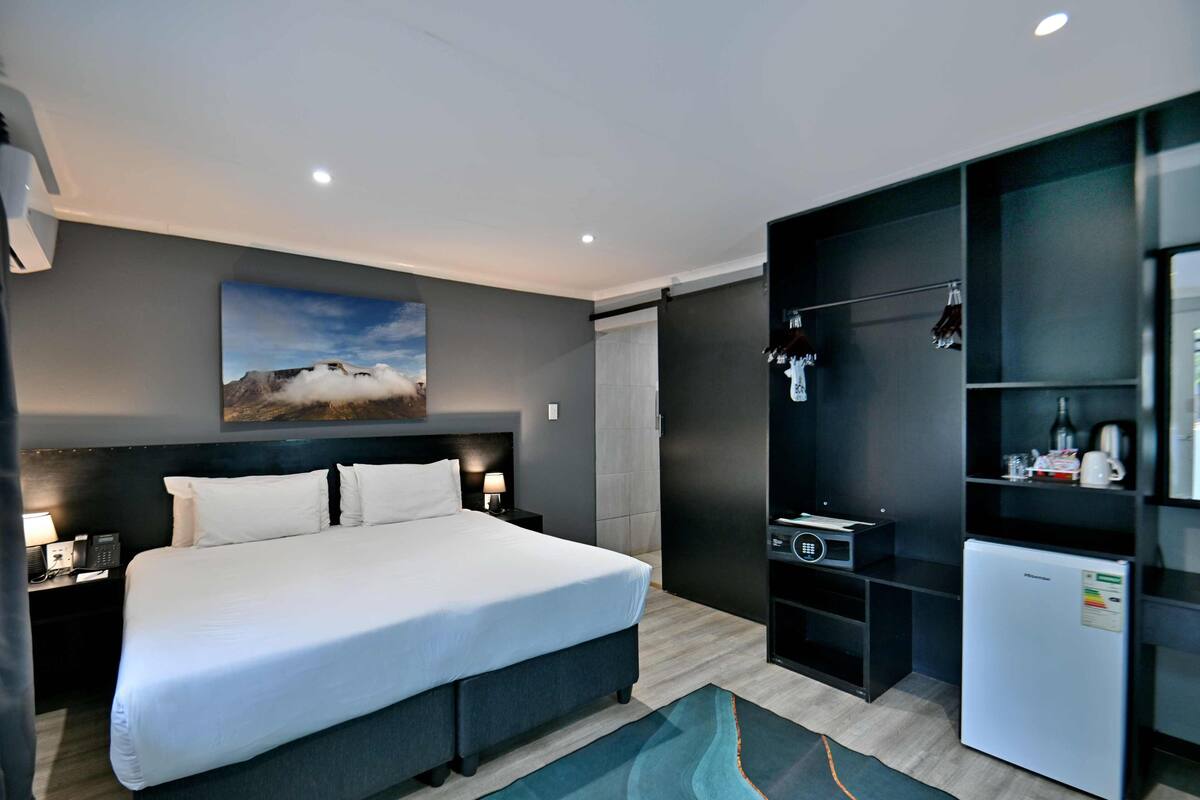 A modern hotel room with a large bed, bedside tables, a small refrigerator, and a built-in TV.