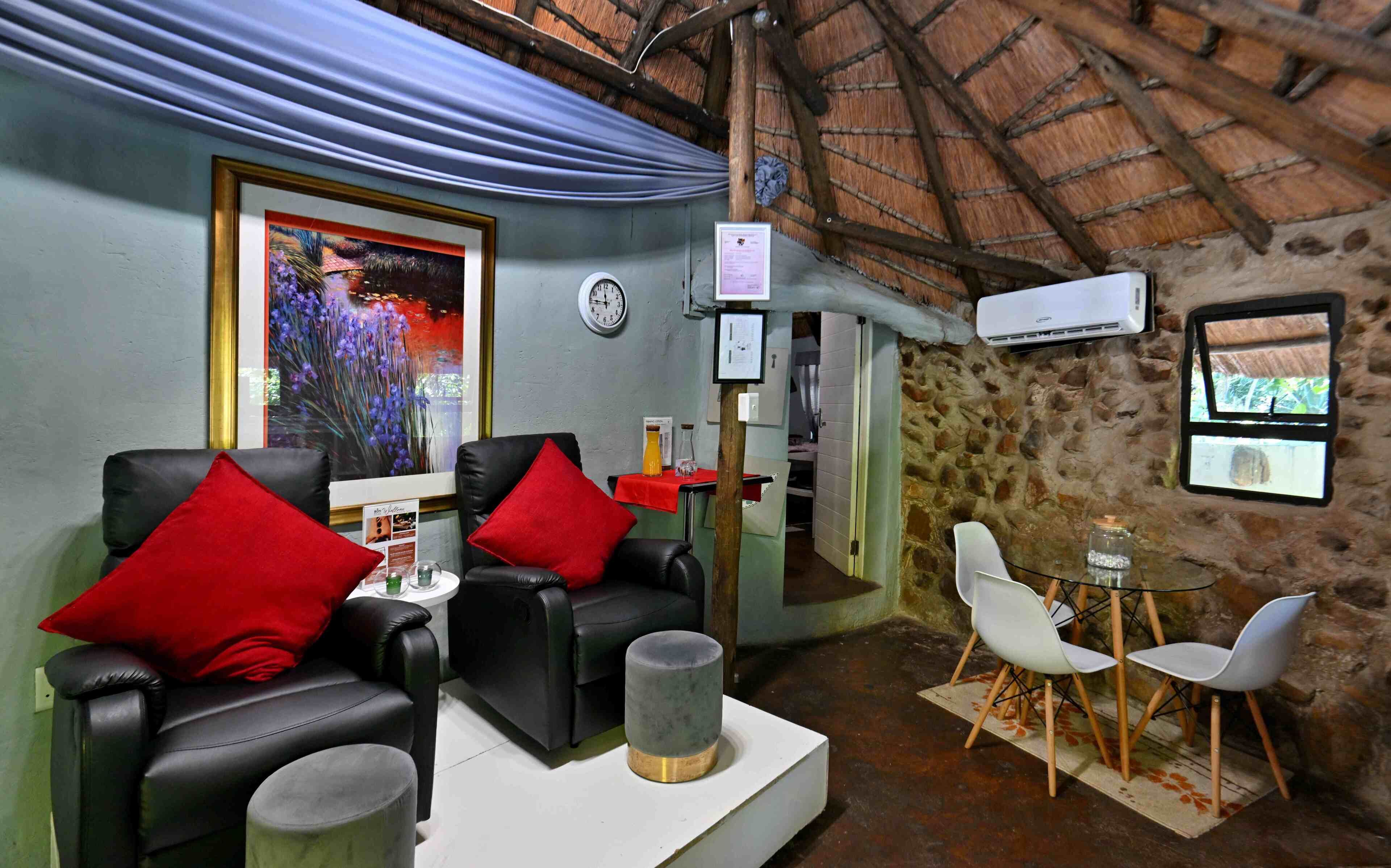 A room with a stone wall, a thatched roof, a clock, a framed painting, and a small table with chairs.