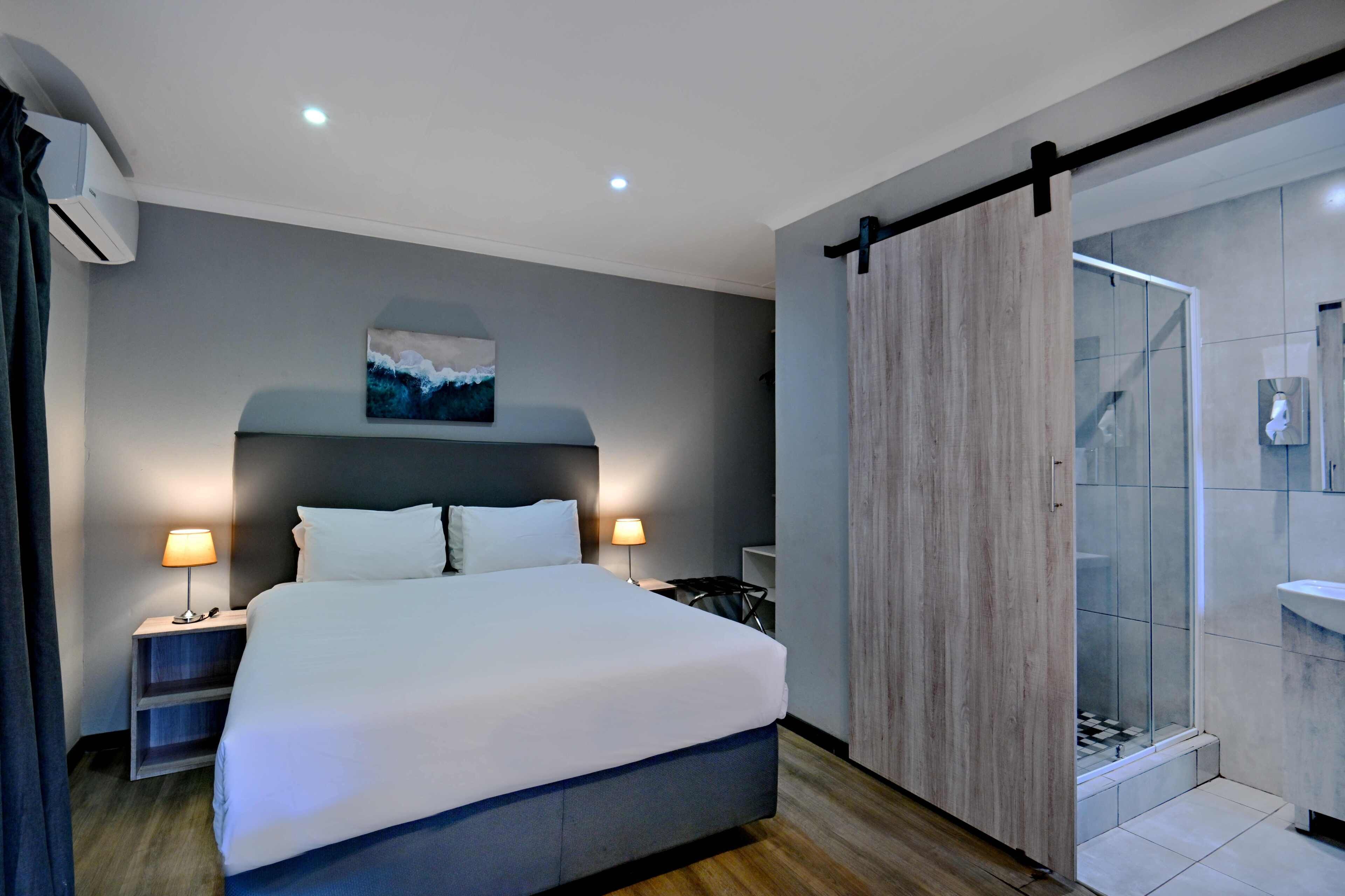 Une chambre d’hôtel moderne avec un grand lit, des lampes de chevet, un tableau au mur, une porte coulissante donnant sur une cabine de douche et un appareil de climatisation visible.