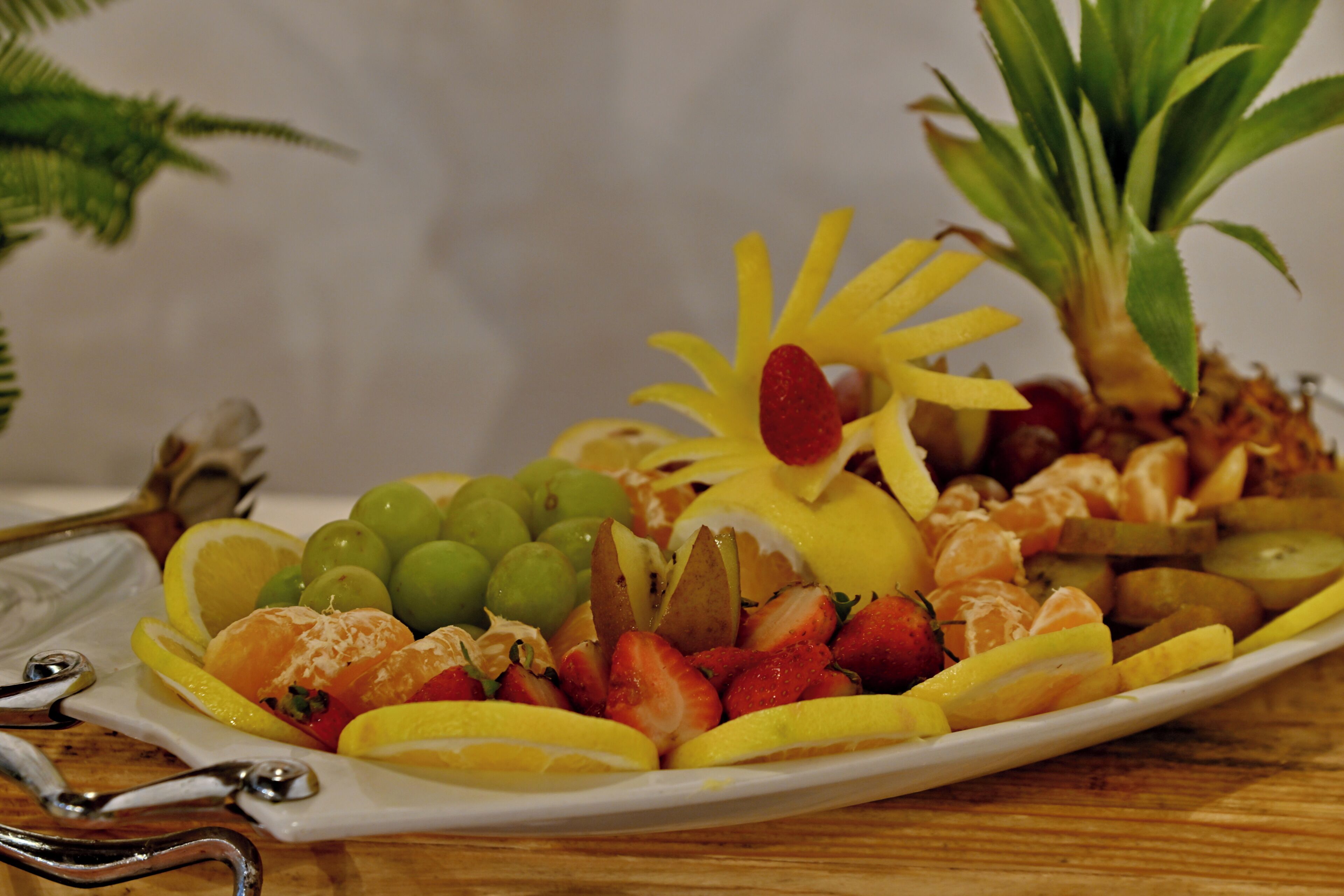 Una bandeja de frutas con diversas frutas y una flor decorativa sobre un plato blanco.