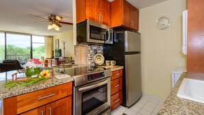 A modern kitchen with wooden cabinets, stainless steel appliances, a granite countertop, and a view of the outdoors. - Top Floor Ocean View & easy walk to the beach. Last minute booking 20 % discount (Kihei)