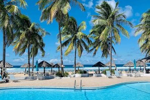 A poolside area with lounge chairs and umbrellas, palm trees, and a view of the ocean. - Sunset Reef: An Oceanfront 3 Bedroom Condo in Gentle Winds (Christiansted)