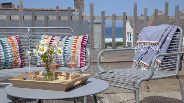 Eine Terrasse mit Metallstühlen und einem Tisch, ein Holztablett mit Speisen und Getränken und Blick aufs Meer durch einen Holzzaun.