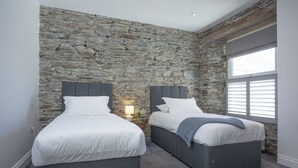 Ein Schlafzimmer mit zwei Betten an einer Steinwand, einem Fenster mit Jalousien und einem Nachttisch mit Lampe.