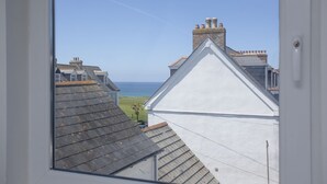 Blick durch ein Fenster eines Gebäudes mit weißem Dach und Schornstein auf eine Küstenlandschaft mit Häusern und dem Meer.