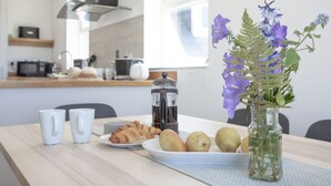 Ein Frühstückstisch mit einem Croissant, einer Obstplatte, einer Kaffeemaschine und einer Vase mit Blumen.