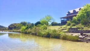 A riverside property with a house overlooking the water, surrounded by greenery and a clear sky. - 4 Maples on the Frio, Riverfront, Wi-Fi, @ Frio River Vacation Rentals (Concan)