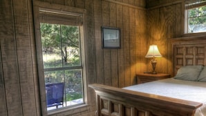 Une chambre à coucher aux murs lambrissés, avec un lit, une table de chevet, une lampe et une fenêtre donnant sur des arbres.