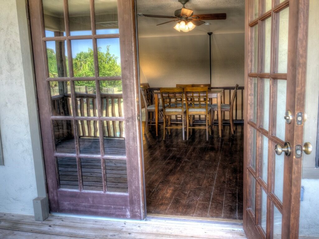 An open wooden door leading to a room with a ceiling fan, a wooden floor, and a view of a wooden deck outside.