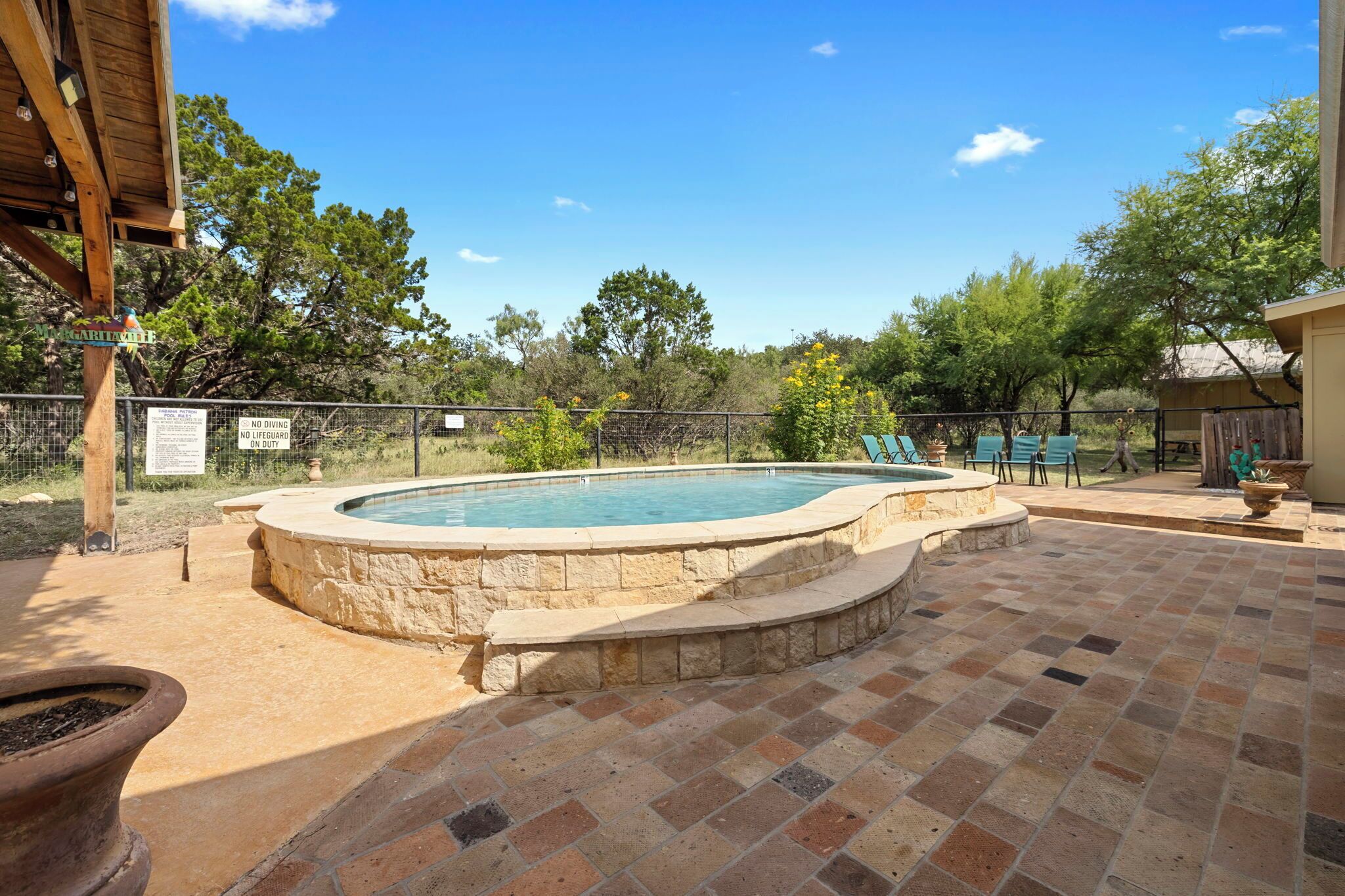A swimming pool with a stone edge, surrounded by a brick patio, and a fenced area with trees and a sign.