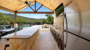 A covered outdoor kitchen with a stone countertop, a stainless steel refrigerator, and a view of a pool and mountains. - Celebration Station, Private Pool, Wi-Fi, @ Frio River Vacation Rentals (Concan)