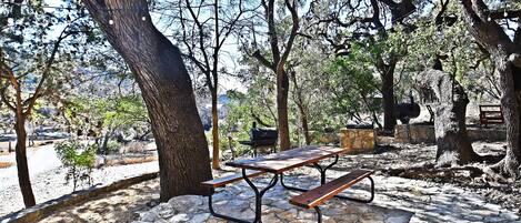 A stone-paved picnic area with a wooden picnic table and benches, surrounded by trees.