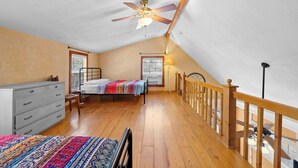 A bedroom with a sloped ceiling, wooden flooring, a bed with a colorful blanket, a dresser, and a ceiling fan. - Stonehaven @ Frio River Vacation Rentals (Concan)