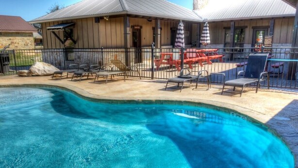 A swimming pool with lounge chairs and umbrellas, surrounded by a fenced patio area with a building in the background. - Rustic Tavern @ Frio River Vacation Rentals (Concan)