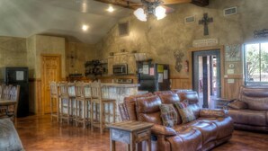 A spacious living room with a leather sofa, wooden dining table, and a ceiling fan. - Rustic Tavern @ Frio River Vacation Rentals (Concan)