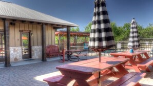 An outdoor patio with picnic tables and striped umbrellas, a covered seating area, and a building with a metal roof. - Rustic Tavern @ Frio River Vacation Rentals (Concan)