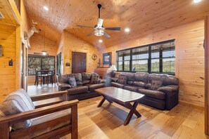 Interior acogedor de una cabaña con techo de madera, ventilador de techo, sofá de cuero, mesa de centro de madera y comedor con mesa y sillas.