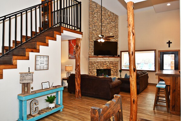 A spacious living room with a stone fireplace, a wooden staircase, and a large TV mounted on the wall.