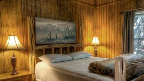 A wooden-paneled room with a bed, two bedside lamps, and a painting on the wall. - Deer Trail @ Frio River Vacation Rentals (Concan)