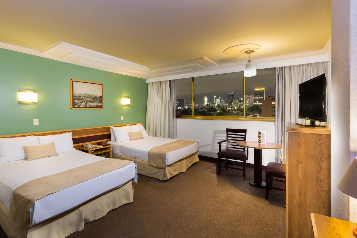 Habitación de hotel con dos camas, un escritorio con silla, televisión y ventana con vista a la ciudad.