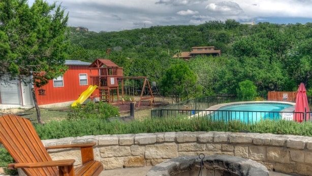 Un jardin avec un foyer en pierre, une balançoire en bois, une maison de jeu rouge et une piscine.