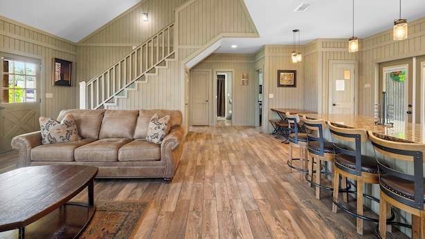 A spacious living area with a high ceiling, a staircase, a wooden floor, a sofa, a coffee table, and a bar area with stools. - Firefly @ Frio River Vacation Rentals (Concan)
