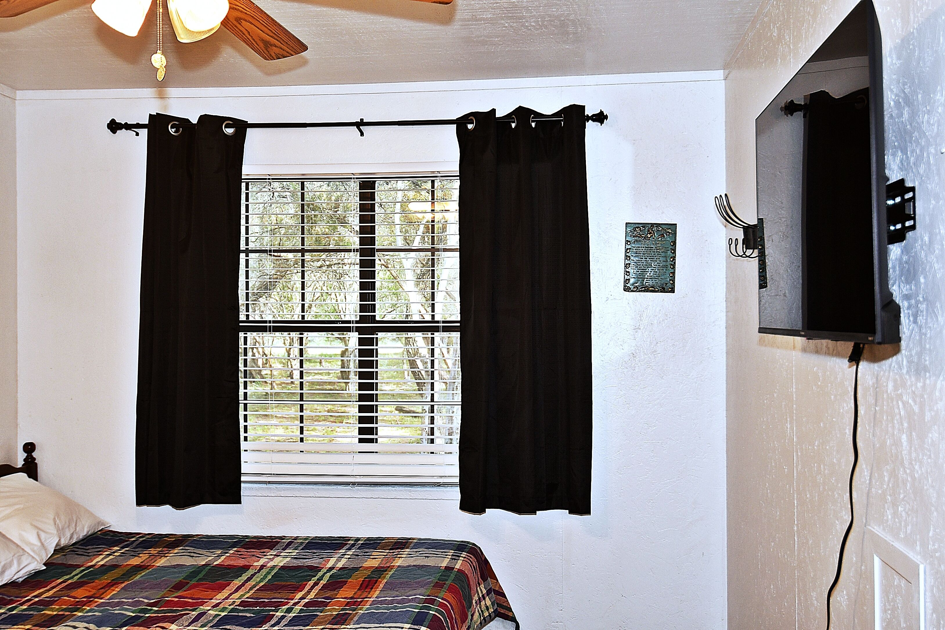 A bedroom with a bed, a window with curtains, a ceiling fan, and a wall-mounted TV.