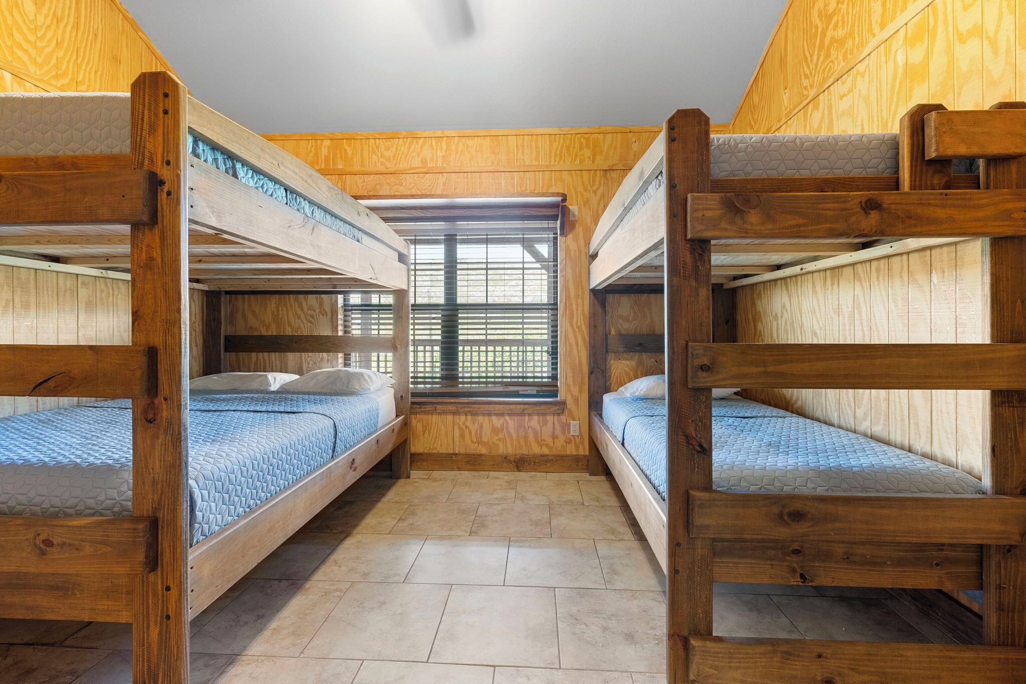 Une pièce avec deux lits superposés, des murs en bois, une fenêtre avec des stores et un sol carrelé.