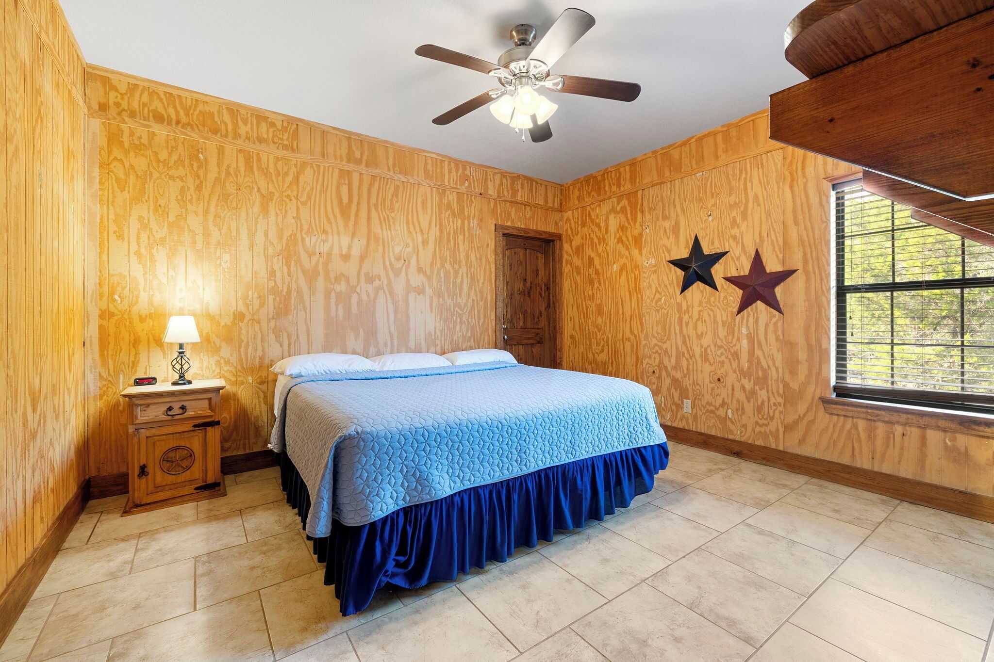 A bedroom with a wooden wall panel, a bed with a blue bedspread, a nightstand with a lamp, a ceiling fan, and a window with blinds.