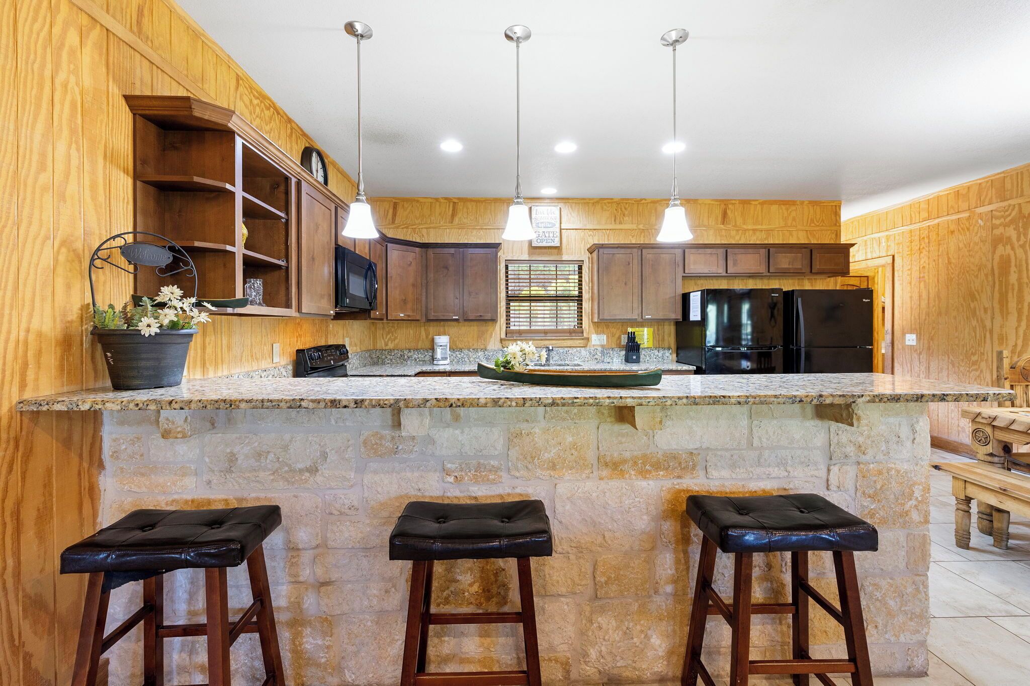 A kitchen with wooden cabinets, a granite countertop, a black refrigerator, and three bar stools.