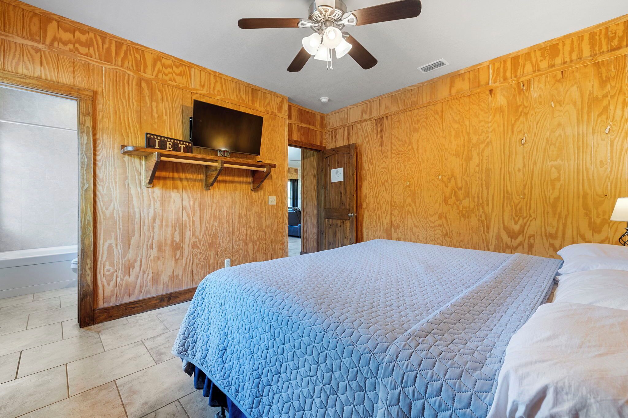 A bedroom with wooden paneling, a bed with a blue quilt, a ceiling fan, and a mounted TV.