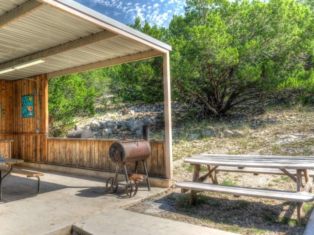 A covered outdoor area with a wooden structure, a barbecue grill, and picnic tables.