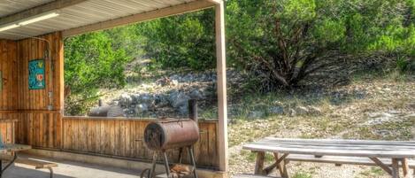 A covered outdoor area with a wooden structure, a barbecue grill, and picnic tables.