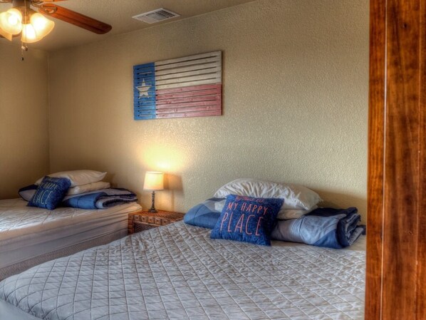 A bedroom with two beds, a ceiling fan, a wall-mounted flag, and a nightstand with a lamp. - Hawk's Ridge @ Frio River Vacation Rentals (Concan)