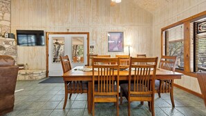 A dining area with a wooden table and chairs, a ceiling fan, a television, and a window with blinds. - Paloma Linda @ Frio River Vacation Rentals (Concan)