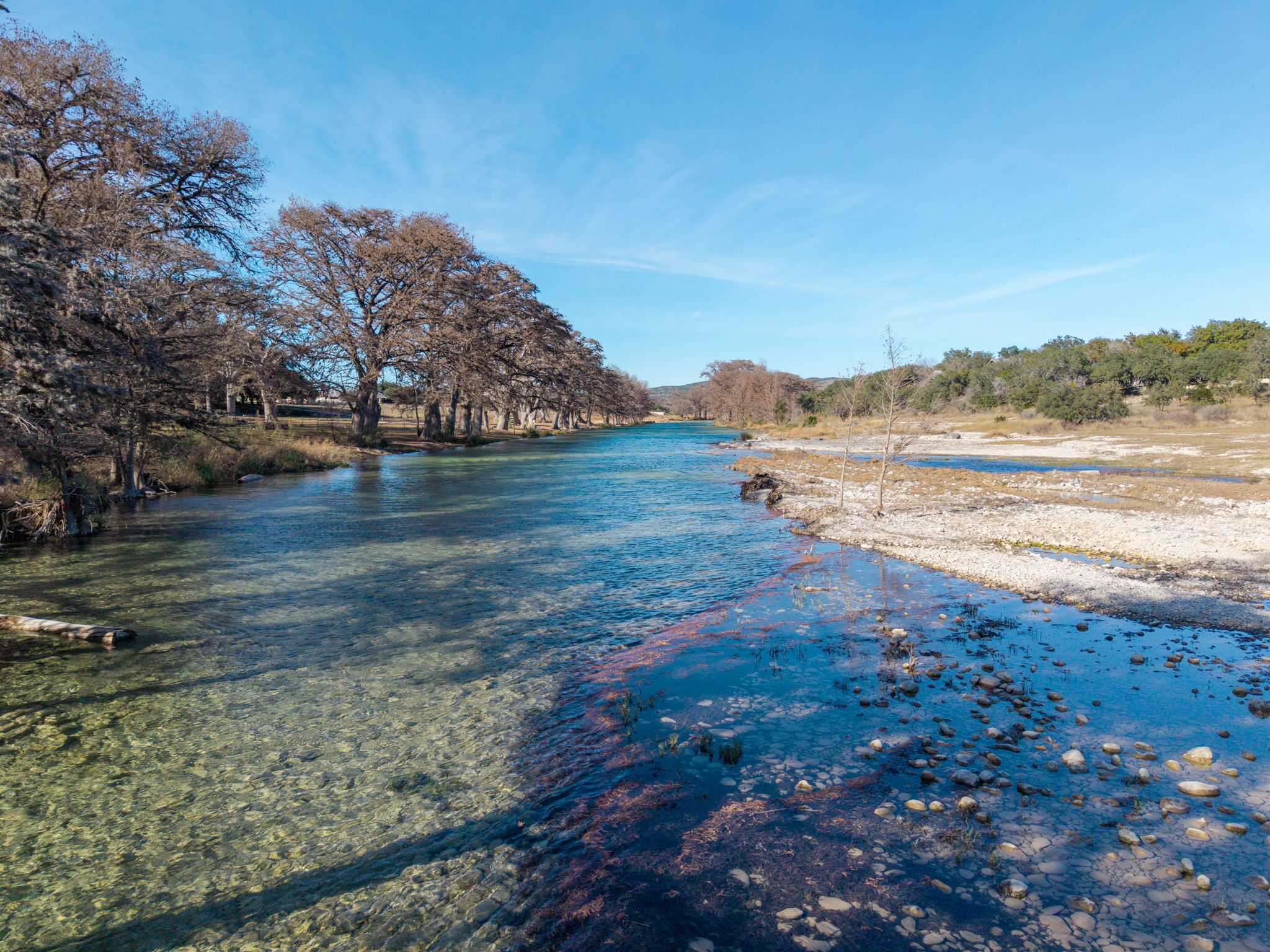A river with clear water, surrounded by trees and a dry landscape.
