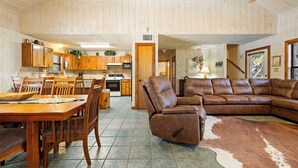 A spacious living room with a brown leather sofa, wooden dining table, and chairs. A kitchen with wooden cabinets and a dining area is visible in the background. - Paloma Linda @ Frio River Vacation Rentals (Concan)