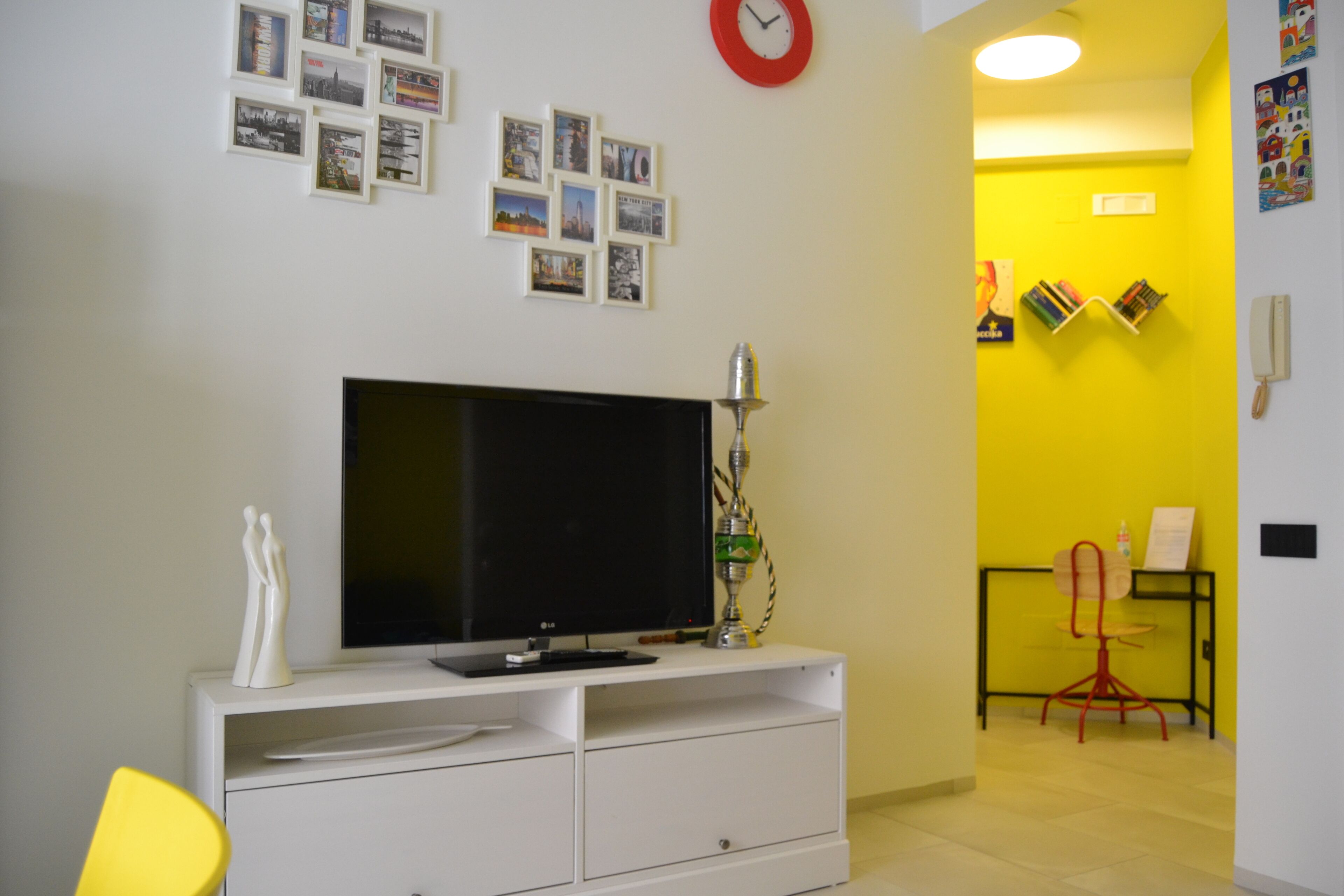 Una sala de estar con un televisor de pantalla plana sobre un mueble blanco, un reloj de pared rojo y una pared amarilla con una estantería y un sillón rojo.