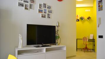 Una sala de estar con un televisor de pantalla plana sobre un mueble blanco, un reloj de pared rojo y una pared amarilla con una estantería y un sillón rojo.