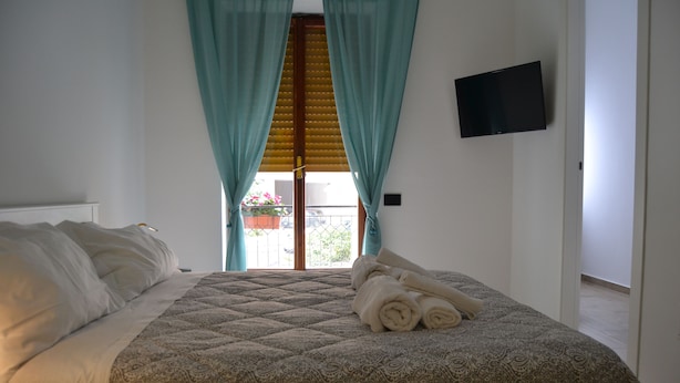 A neatly made bed with a patterned bedspread and rolled towels, a television mounted on the wall, and a view of the outside through a window with curtains. - Alleria Holiday Home (Salerno)