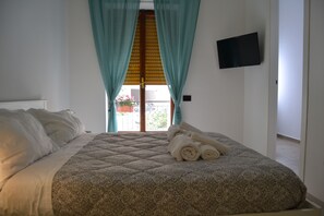 Ein ordentlich gemachtes Bett mit einem gemusterten Bettüberwurf und aufgerollten Handtüchern, ein an der Wand montierter Fernseher und ein Blick ins Freie durch ein mit Vorhängen versehenes Fenster.