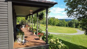 Eine Holzveranda mit Topfpflanzen und einem Tisch, mit Blick auf eine grüne Landschaft mit Bäumen und einem Weg.