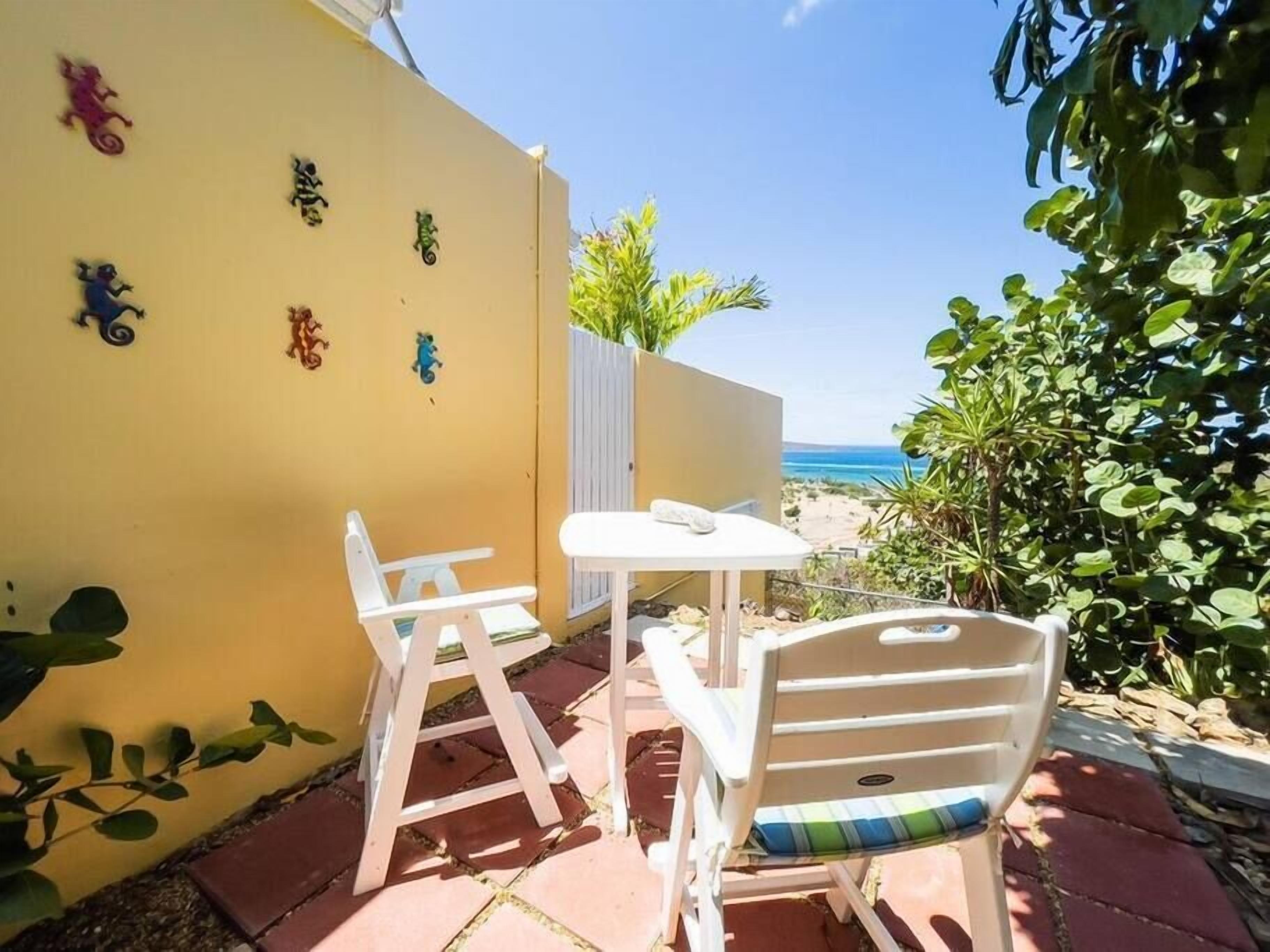 Une terrasse avec une table blanche et deux chaises blanches, un mur jaune orné de décorations murales, et une vue sur la plage.