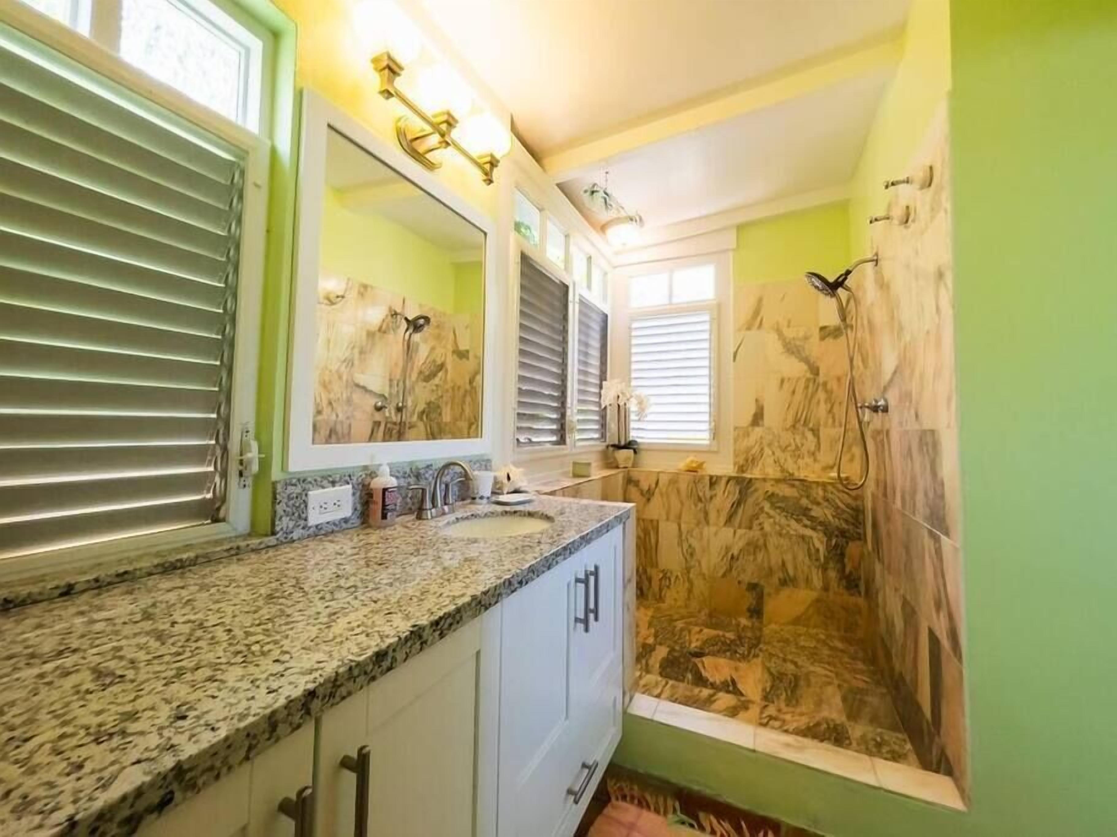 A bathroom with a granite countertop, a large mirror, a shower with a marble wall, and a window with blinds.