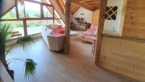 A cozy loft bedroom with a sloped ceiling, wooden beams, a large window, a white sofa, a red patterned bed, and a TV. - Le Typique Atypique Spacieux 140 m2 au Centre du Hameau de Perroix (talloires montmin)